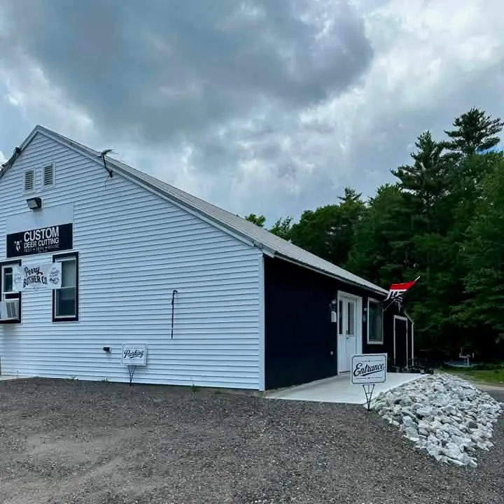 The Perry Butcher Co. shop in East Waterboro, Maine — a white gable-end building with the Perry Butcher Co. and Custom Deer Cutting signs, an Entrance sign by the door, and the gravel parking area in front